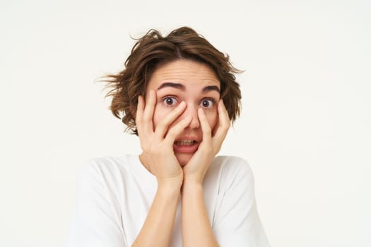 Image of girl screams and looks scared, hides face with hands, stands over white background. Copy space