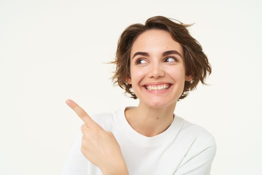 Image of cheerful brunette female model, candid girl points finger left and smiles, shows advertisement, poses over white background.