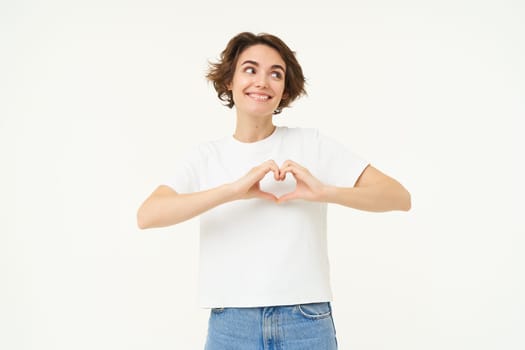 Portrait of brunette girl shows heart sign, love gesture, express care and warm feelings, stands over white background.