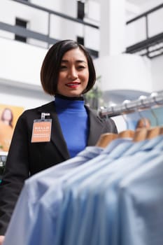 Smiling asian woman assistant with badge looking at apparel hanging on rack while working in clothing store. Shopping center boutique cheerful employee managing merchandise stock