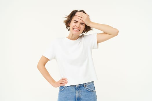 Health and people. Young woman has headache, touches her head and grimacing from painful migraine, feeling unwell, dizzy, standing over white background.