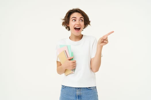 Portrait of woman, student with amazed face, carry her notebooks and documents, points at upper right corner, shows advertisement, studio background.