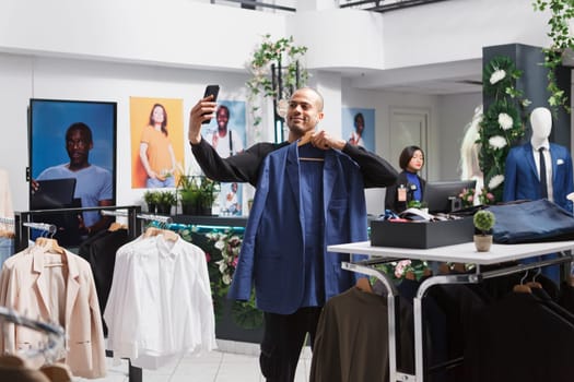 Young arab man internet influencer live streaming using smartphone in clothing store while showcasing jacket. Boutique client choosing outfit and showing apparel on hanger on mobile phone front camera