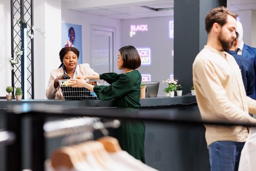 Black Friday and retail industry. Young Asian woman customer standing at clothing store checkout counter giving clothes to friendly cashier, buying garment at discounted prices during seasonal sales