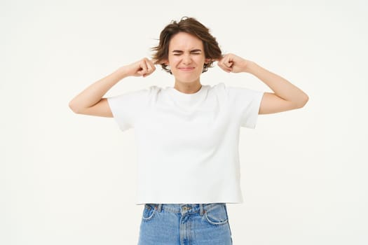 Woman standing annoyed, holding fingers in ears, unwilling to listen, feeling discomfort from loud noise, standing over white background.