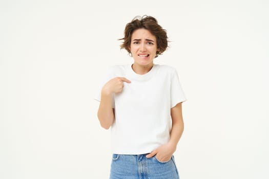 Portrait of awkward girl, feeling uncomfortable, pointing at herself with unsure face, posing over white background.