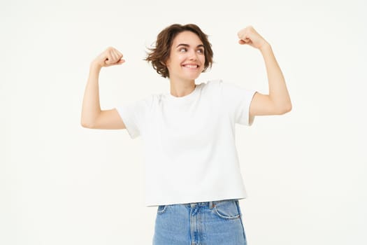 Young happy girl feeling strong, woman shows biceps, flexing muscles on arms and smiling, proves her strength, stands over white background.