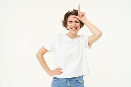 Portrait of happy young woman mocking, winning prize and making fun of loser, showing l letter on forehead, stands over white background.