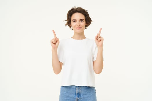 Image of happy, beautiful modern woman, pointing fingers up, showing advertisement on top, smiling, standing over white background.