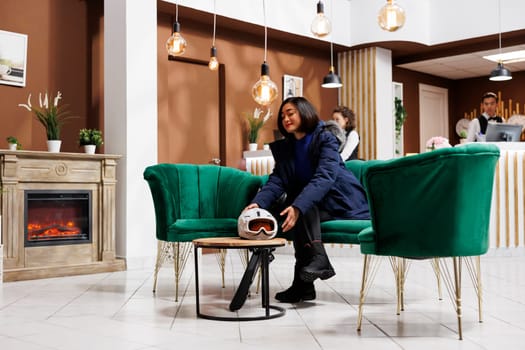 Young female tourist sitting on sofa in lounge area of luxurious winter resort waiting for accommodation. Enthusiastic woman placing ski helmet on table of hotel lobby ready for wintertime activities.