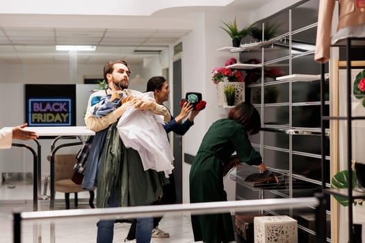 Stressed Black Friday obsessed diverse people running with clothes around fashion store with empty shelves, customers hunting for bargains during big seasonal sales in shopping mall