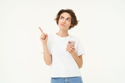 Portrait of woman placing an order online on smartphone app, pointing at upper left corner and looking with thinking face, making decision, standing thoughtful over white background.