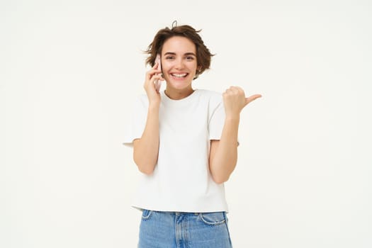 Portrait of friendly, smiling cute girl, pointing finger right, talking on mobile phone, showing smth aside on white copy space, isolated on studio background.