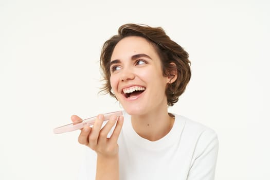 Image of woman records voice message on smartphone, talking in speakerphone, stands over white background.