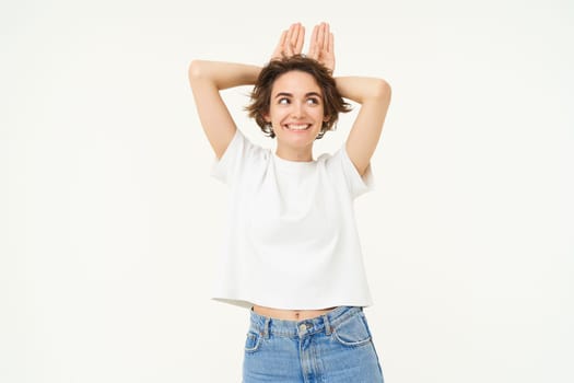 Portrait of funny and cute young woman with bunny ears gesture, holding palms on top of her head, smiling and looking happy at camera, standing over white background.