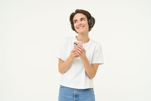 Image of happy young girl listens to music in headphones, standing with smartphone, using mobile phone streaming app, white background.