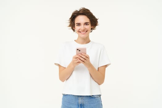 Portrait of brunette woman using smartphone, standing with mobile phone, texting, placing an order, browsing social media in telephone, standing over white background.