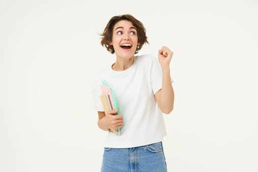 Enthusiastic brunette woman makes fist pump, holds documents and notes, looks thrilled and happy, winning, triumphing, posing over white background.