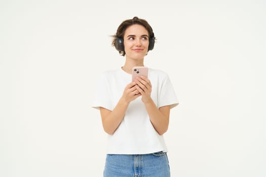 Portrait of smiling brunette woman with smartphone, smiling, looking aside, listening to music, standing over white studio background.