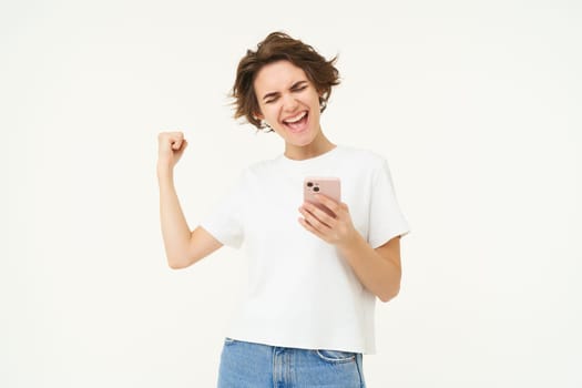 Portrait of excited brunette woman, holding smartphone and celebrating big win, achieves goal in mobile app, stands over white background. Copy space