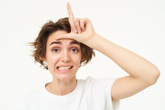 Close up of young woman making fun of friend, smiling and showing loser gesture, l letter on forehead, standing over white background.