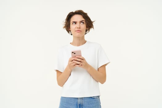 Portrait of thinking woman with smartphone, looking puzzled up, pondering, making decision, standing against white studio background. Copy space