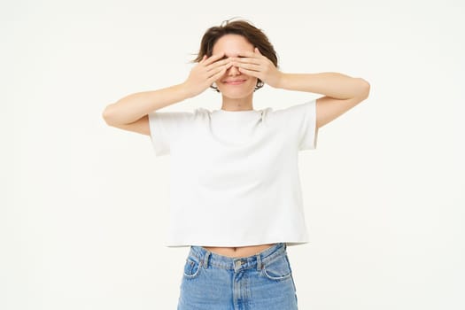 Portrait of happy brunette girl, stands with closed eyes, blindsided, waits for surprise, stands over white background. Copy space