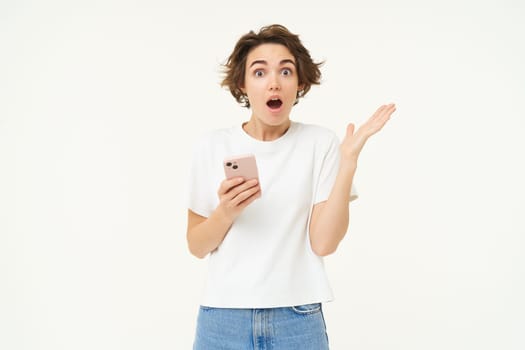 Portrait of brunette girl with shocked face, reading something amazing on mobile phone, standing with smartphone over white background.