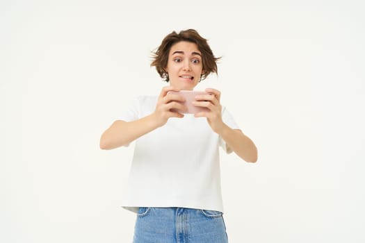 Portrait of woman playing video games on mobile phone, holding smartphone with both hands, standing over white studio background.