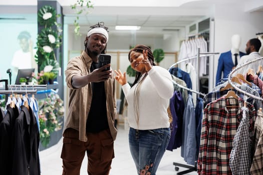 African american man and woman influencers taking selfie in fashion boutique. Smiling social media bloggers posing for mobile phone photo while promoting clothing brand in shopping mall