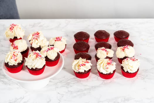 Piping white chocolate ganache frosting on top of red velvet cupcakes and topping with sprinkles.