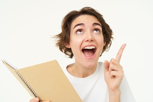 Enthusiastic brunette woman with diary, holding planner book, pointing at upper right corner and smiling with excitement, standing over white background.