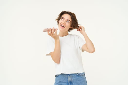 Portrait of chatty woman talking in speakerphone, records voice message on mobile phone and smiling, stands over white background.