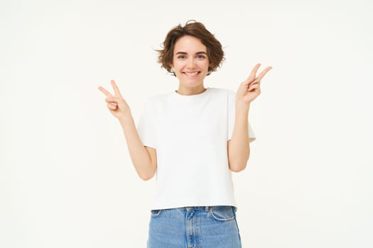 Image of happy, young positive woman, shows v-sign, peace gestures and smiling, standing over white studio background.