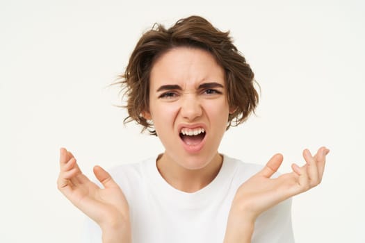 Close up portrait of brunette woman raising hands up and frowning, looking angry, shocked and offended, shouting, isolated over white background.