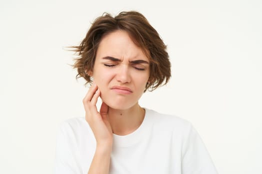Close up of woman has a toothache, touches her teeth and frowns from painful discomfort, stands over white background.