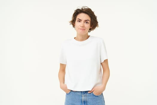 Portrait of woman looking reluctant and unconfident, frowning, holding hands in pockets, standing doubtful, feeling unsure, white studio background.