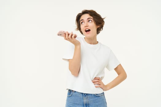 Portrait of chatty woman talking in speakerphone, records voice message on mobile phone and smiling, stands over white background.