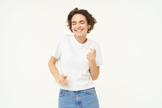 Portrait of carefree, happy young woman in wireless headphones, listens to music, dances to favourite song in earphones, posing over white studio background.