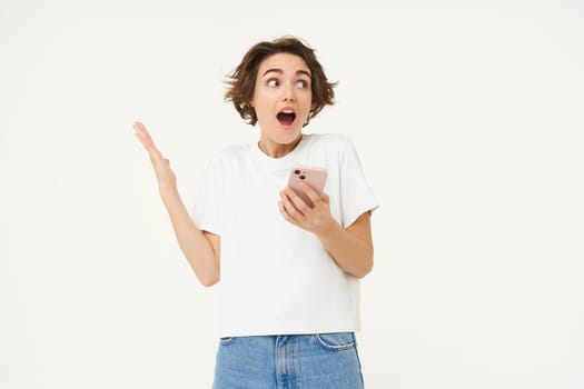Portrait of brunette woman with smartphone, looking surprised and amazed, checking out big news on mobile phone, standing excited against white background.