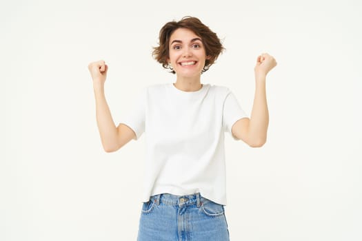 Excited brunette girl, student winning prize, celebrating victory, triumphing, standing in white t-shirt and jeans over white background.