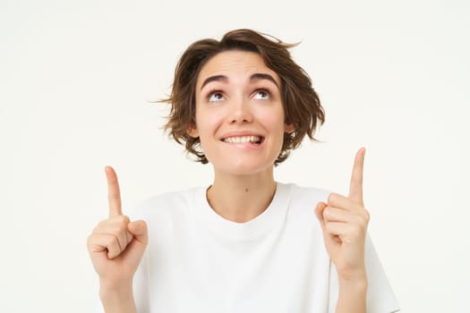 Portrait of happy, beautiful young woman pointing fingers up, showing advertisement or product on top of copy space, standing against white background.