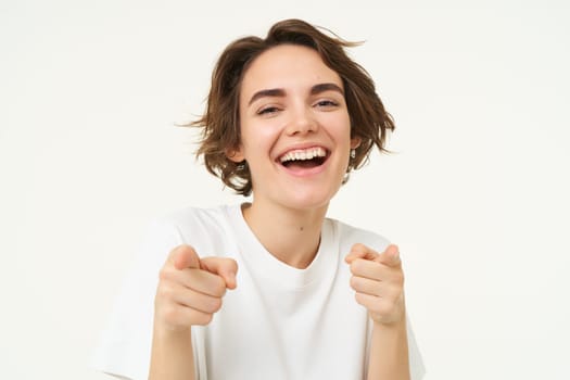 Close up portrait of cute smiling woman, pointing fingers at camera, choosing, inviting you, posing against white studio background.