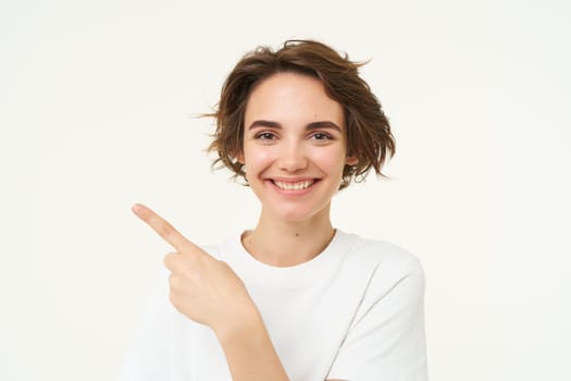 Close up shot of happy, candid woman, laughing and smiling, pointing finger left, showing advertisement, standing over white background.