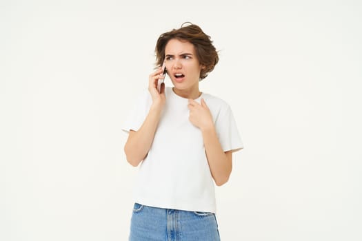 Portrait of shocked, angry woman talks on mobile phone with disappointed, furious face expression, standing over white background.