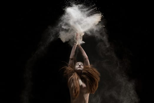 Slim young barefoot woman throwing sand over head while dancing against black background