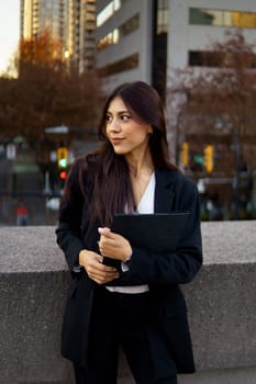 Professional businesswoman standing on a sidewalk in front of a modern building with autumn foliage.