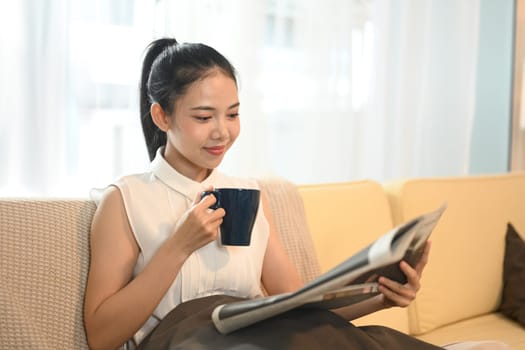 Beautiful young woman enjoys her morning cup and reading a newspaper in living room.