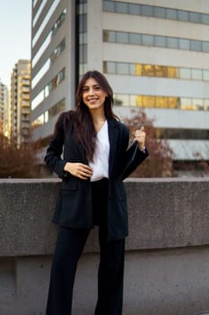 Professional businesswoman standing on a sidewalk in front of a modern building with autumn foliage.