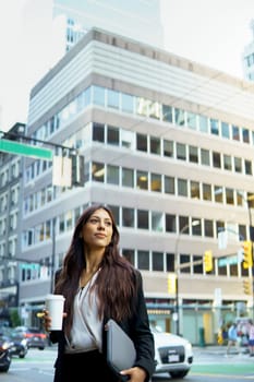 Young businesswoman in black suit holding a coffee cup walking on a busy city street.